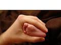 Stambiu planu – suaugusio žmogaus ranka švelniai laikanti mažytę kūdikiui pėdelę, primenančią raminamąjį masažą, švelniu, tamsiu fonu.