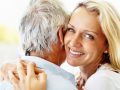 Moteris ilgais šviesiais plaukais šiltai šypsosi prieš kamerą apsikabindama vyresnio amžiaus vyrą žilais plaukais, įkūnijantį sveikas senėjimo esmę. Nustatymas atrodo šviesus ir džiaugsmingas.