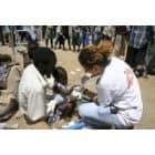 Baltu chalatu vilkintis sveikatos priežiūros darbuotojas klūpo ir apžiūrinėja ant moters kelių sėdintį mažą vaiką. Aplink juos susirenka minia žmonių lauko, galbūt reljefo stovykloje.