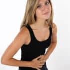 Asmuo su ilgais plaukais, dėvintis juodą viršutinę dalį, sulaiko pilvą, galbūt dėl endometriozės, stovėdamas baltame fone.