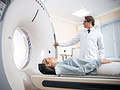 Pacientas guli ant skenuojančios lovos, įeidamas į kompiuterinės tomografijos aparatą, o gydytojas baltu chalatu koreguoja nustatymus naudodamas apšvitos optimizavimą. Patalpa yra gerai apšviesta ir šiuolaikiška, gydytojas sutelkė dėmesį į efektyvų įrangos valdymą.