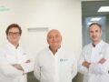 Trys gydytojai baltais chalatais stovi moderniame medicinos kabinete ir šiltai šypsosi. Ant sienos matomas logotipas "Clinic Agatas", garsėjantis profesionaliomis stuburo operacijomis ir mikrodiskektomijos procedūromis.